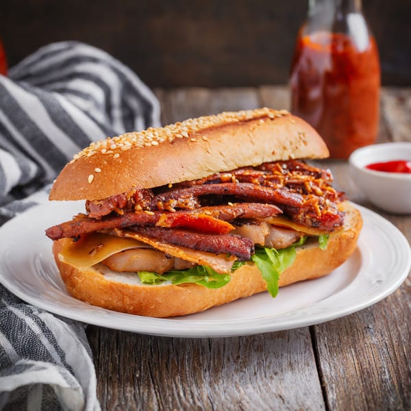 A soul food sandwich with meat, cheese, and lettuce on a sesame seed bun, with hot sauce.