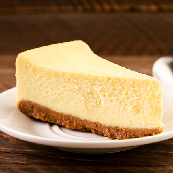 A slice of classic cheesecake with a graham cracker crust on a white plate.