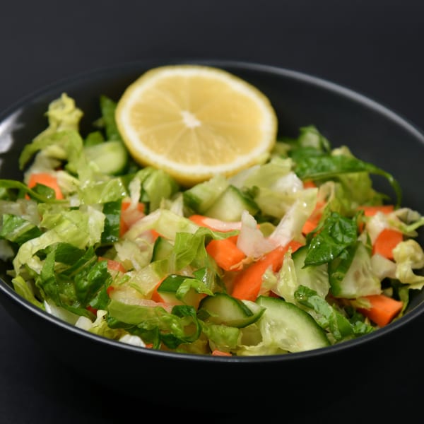 Fresh side salad with lettuce, cucumbers, carrots, and a lemon wedge in a black bowl.